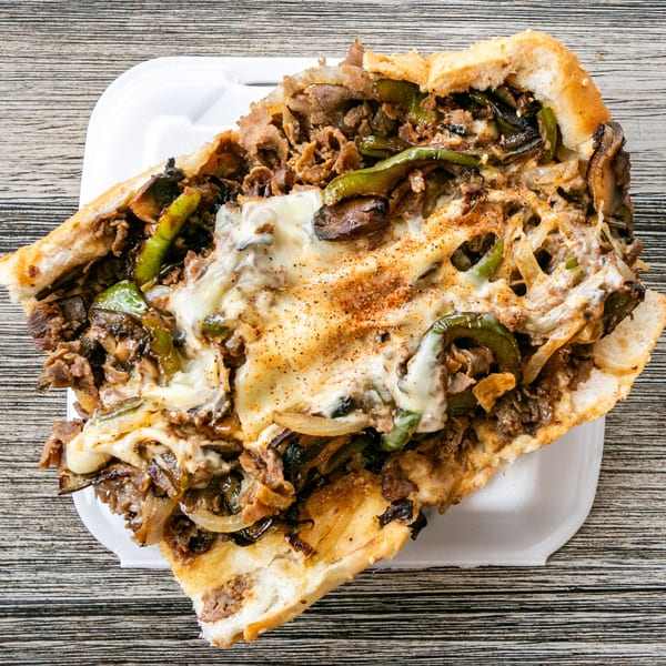 Cheesesteak sandwich with steak, melted cheese, green peppers, onions, and mushrooms in a white container.
